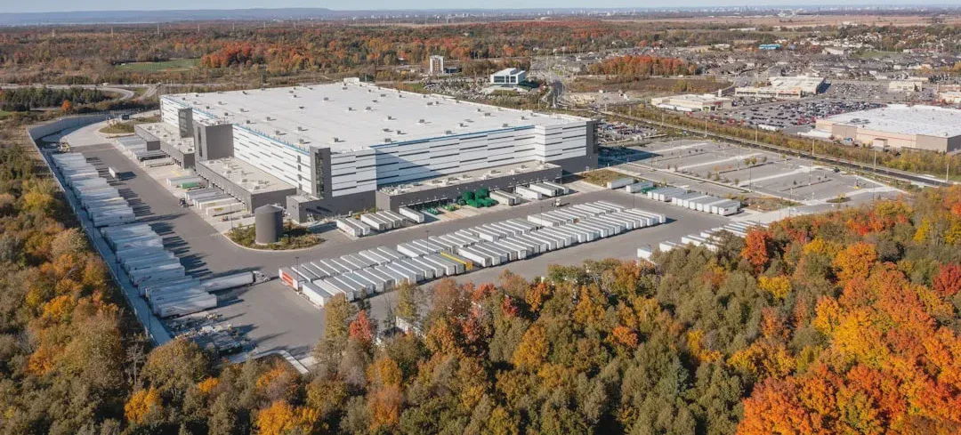 Zdjęcie przedstawia duży, nowoczesny kompleks magazynowy otoczony jesienną zielenią. W scenerii widoczne są ciężarówki z oznaczeniami Amazon, co sugeruje centrum realizacji zamówień lub węzeł logistyczny. — Zdjęcie: Point3D Commercial Imaging Ltd.